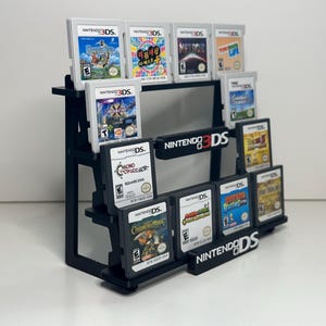 May include: A black tiered display stand holding multiple Nintendo 3DS and DS game cartridges. The stand features the text NINTENDO 3DS on the middle tier and NINTENDO DS on the bottom tier. The cartridges are arranged in rows on the black shelves.