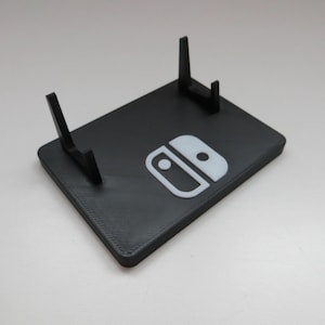 May include: A black plastic stand with two supports for a Nintendo Switch console. The stand has a white Nintendo Switch logo on the front.