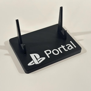 Peut inclure: Support noir avec deux supports verticaux et une base rectangulaire. La base porte le mot "Portal" en blanc, ainsi qu'un logo PlayStation blanc. Le support est conçu pour maintenir un appareil de jeu.