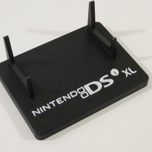 May include: Black plastic stand for a Nintendo DS XL. The stand has two vertical supports and the words "NINTENDO DS XL" printed in white.