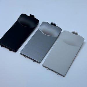 Peut inclure: Trois couvercles de batterie de remplacement pour un appareil de jeu portable. Les couvercles sont noirs, argentés et blancs.