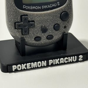 Peut inclure: Un support noir imprimé en 3D tenant un appareil Pokémon Pikachu 2 argenté et noir. Le support porte les mots "POKÉMON PIKACHU 2" en lettres blanches. L'appareil possède un pavé directionnel et des boutons.