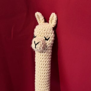 May include: A crocheted llama head with closed eyes is attached to the handle of a white and red tennis racket. The racket is against a red background.