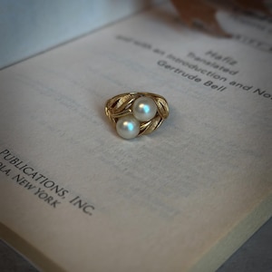 May include: A gold ring with two white pearls, set in a leaf design. The ring is resting on an open book with the text "Hafiz Translated as introduction and No Gertrude Bell PUBLICATIONS, INC. LA, NEW YORK."