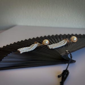 May include: Elegant pearl earrings with gold accents rest on a black folding fan. The earrings feature a large pearl stud and a cascade of smaller pearls. The fan is open, displaying its pleated design, and is set against a white background.