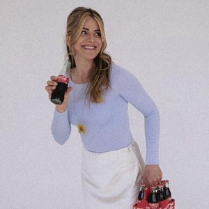 May include: A woman holding a Coca-Cola bottle and a six-pack of Coca-Cola bottles. She is wearing a light blue long-sleeve shirt and a white skirt. The Coca-Cola bottles are in a red carrier.