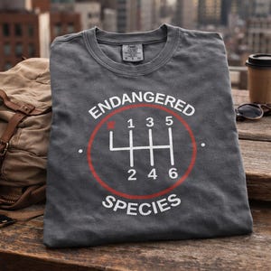 May include: A charcoal grey t-shirt featuring a white graphic of a six-speed manual transmission gear shift pattern enclosed in a red circle. The text ENDANGERED is printed above the circle and SPECIES is printed below in white capital letters.