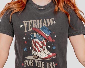 Può includere: T-shirt grigio scuro con una grafica di stivali da cowboy rossi, bianchi e blu e un cappello abbinato. Il testo recita "YEEHAW" sopra e "FOR THE USA" sotto, con accenti di stelle. La maglietta è etichettata "PEPPER".
