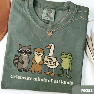 K&ouml;nnte beinhalten: Moosgr&uuml;nes T-Shirt mit einer Grafik eines Waschb&auml;ren, eines Otters, einer Ente mit einem Schild mit der Aufschrift "You Matter" und eines Frosches. Der Text "Celebrate minds of all kinds" ist unter den Tieren aufgedruckt. Eine silberne Halskette liegt auf dem Shirt.