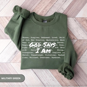 K&ouml;nnte beinhalten: Milit&auml;rgr&uuml;nes Sweatshirt mit wei&szlig;em Text. Der Text enth&auml;lt S&auml;tze wie "Gott sagt, ich bin" und andere inspirierende Worte. Das Sweatshirt wird auf einer hellen Holzmaserungsoberfl&auml;che pr&auml;sentiert.