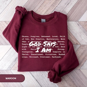 K&ouml;nnte beinhalten: Burgunderfarbenes Sweatshirt mit wei&szlig;em Text, der "GOD SAYS I AM" und eine Liste von Affirmationen zeigt. Das Wort "MAROON" befindet sich in einem rechteckigen Feld unten links. Das Sweatshirt hat einen Rundhalsausschnitt.