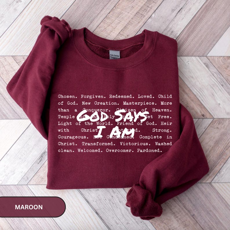 K&ouml;nnte beinhalten: Burgunderfarbenes Sweatshirt mit wei&szlig;em Text, der "GOD SAYS I AM" und eine Liste von Affirmationen zeigt. Das Wort "MAROON" befindet sich in einem rechteckigen Feld unten links. Das Sweatshirt hat einen Rundhalsausschnitt.