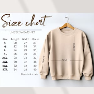 K&ouml;nnte beinhalten: Ein beiges Unisex-Sweatshirt h&auml;ngt an einem Holzb&uuml;gel. Das Bild enth&auml;lt eine Gr&ouml;&szlig;entabelle mit Ma&szlig;en in Zoll f&uuml;r L&auml;nge, Breite und &Auml;rmel f&uuml;r die Gr&ouml;&szlig;en S bis 5XL. Die Tabelle ist mit "UNISEX SWEATSHIRT" beschriftet.