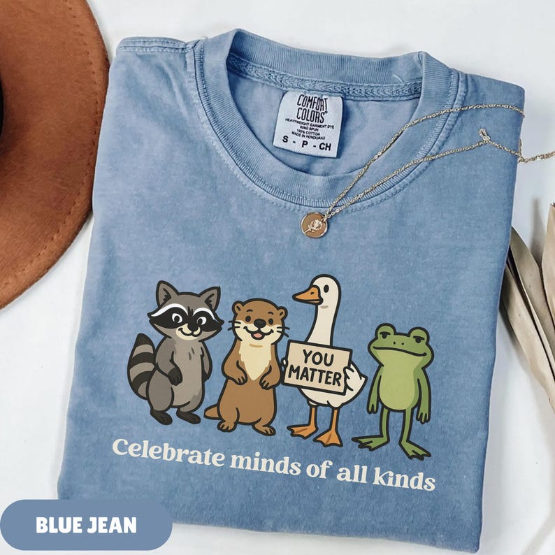 K&ouml;nnte beinhalten: Blaues Jeans-T-Shirt mit einer Grafik eines Waschb&auml;ren, eines Otters, einer Ente mit einem Schild mit der Aufschrift "You Matter" und eines Frosches. Der Text "Celebrate minds of all kinds" ist darunter gedruckt. Eine goldene Halskette liegt auf dem Shirt.