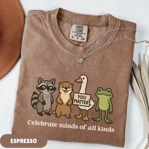 K&ouml;nnte beinhalten: Espressofarbenes T-Shirt mit Rundhalsausschnitt und kurzen &Auml;rmeln. Das Shirt zeigt eine Grafik eines Waschb&auml;ren, eines Otters, einer Ente mit einem Schild mit der Aufschrift "You Matter" und eines Frosches. Der Text "Celebrate minds of all kinds" ist darunter gedruckt.