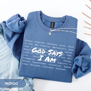 K&ouml;nnte beinhalten: Indigoblaues Sweatshirt mit Rundhalsausschnitt und dem Text "GOD SAYS I AM" sowie einer Liste von Affirmationen in Wei&szlig;. Eine silberne Halskette mit Anh&auml;nger liegt auf dem Shirt. Das Sweatshirt ist auf einer Jeans gefaltet.