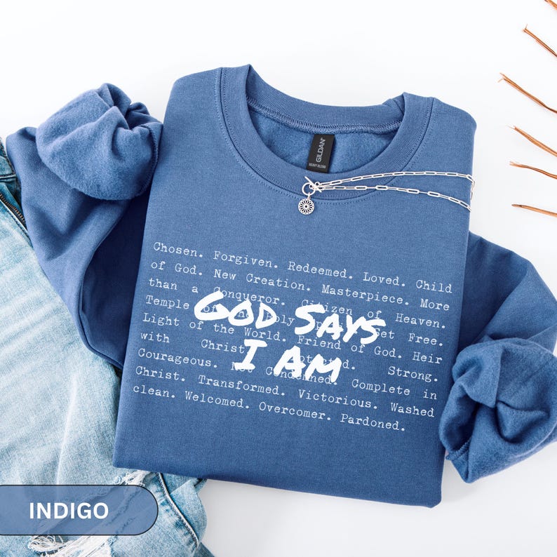 K&ouml;nnte beinhalten: Indigoblaues Sweatshirt mit Rundhalsausschnitt und dem Text "GOD SAYS I AM" sowie einer Liste von Affirmationen in Wei&szlig;. Eine silberne Halskette mit Anh&auml;nger liegt auf dem Shirt. Das Sweatshirt ist auf einer Jeans gefaltet.