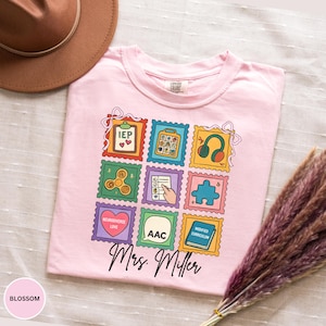 May include: A light pink t-shirt with a colorful stamp design featuring educational symbols and the text "Mrs. Miller." The shirt has a crew neck and short sleeves. The word "Blossom" is printed on the bottom left.