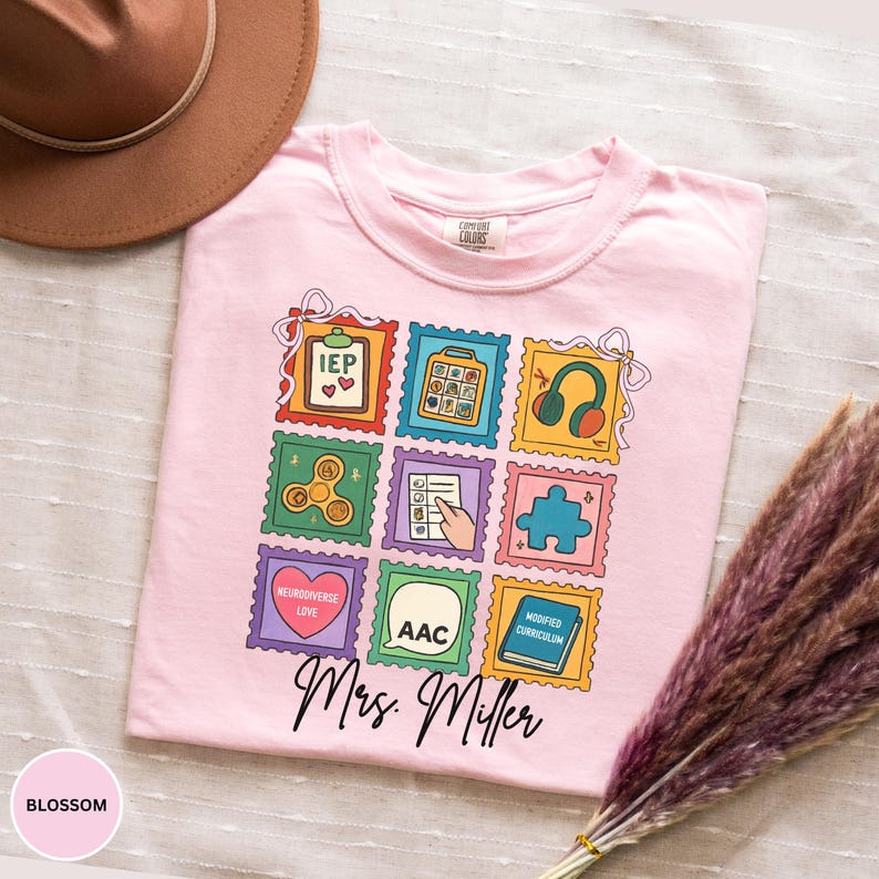 May include: A light pink t-shirt with a colorful stamp design featuring educational symbols and the text "Mrs. Miller." The shirt has a crew neck and short sleeves. The word "Blossom" is printed on the bottom left.