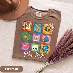 May include: Espresso-colored t-shirt featuring a colorful stamp design with various icons, including headphones, a puzzle piece, and the text "IEP." The shirt also includes the name "Mrs. Miller" in a script font.
