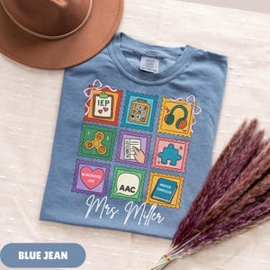 May include: Blue jean t-shirt with colorful stamp-like graphics. The stamps feature images of a calculator, headphones, a puzzle piece, and a fidget spinner. The shirt also has the text "Mrs. Miller" in a script font.