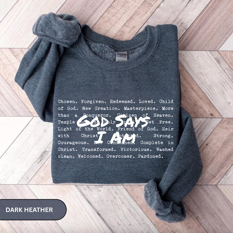 K&ouml;nnte beinhalten: Dunkelgraues Sweatshirt mit wei&szlig;em Text. Der Text enth&auml;lt die S&auml;tze &bdquo;Gott sagt, ich bin&ldquo; und eine Liste von Affirmationen. Das Sweatshirt ist gefaltet, die &Auml;rmel sind &uuml;ber die Oberseite drapiert.