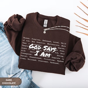 K&ouml;nnte beinhalten: Ein dunkelbraunes Sweatshirt mit den wei&szlig;en Worten "GOD SAYS I AM". Das Sweatshirt hat eine Liste inspirierender W&ouml;rter aufgedruckt. Eine silberne Halskette mit Anh&auml;nger liegt auf dem Sweatshirt.