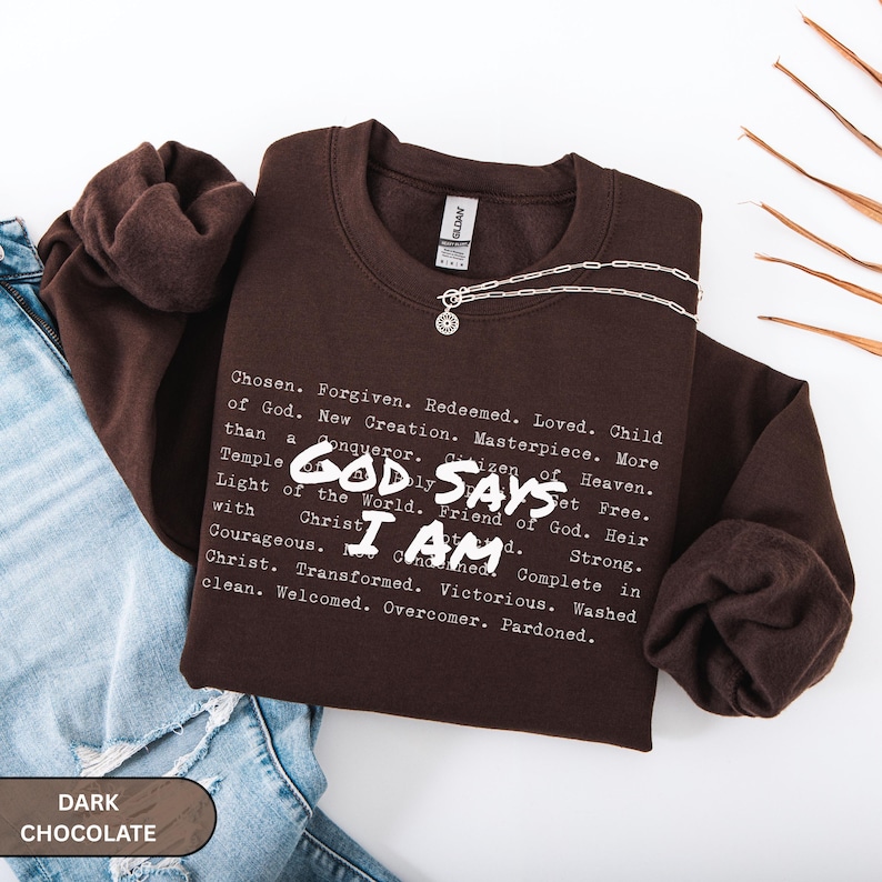 K&ouml;nnte beinhalten: Ein dunkelbraunes Sweatshirt mit den wei&szlig;en Worten "GOD SAYS I AM". Das Sweatshirt hat eine Liste inspirierender W&ouml;rter aufgedruckt. Eine silberne Halskette mit Anh&auml;nger liegt auf dem Sweatshirt.