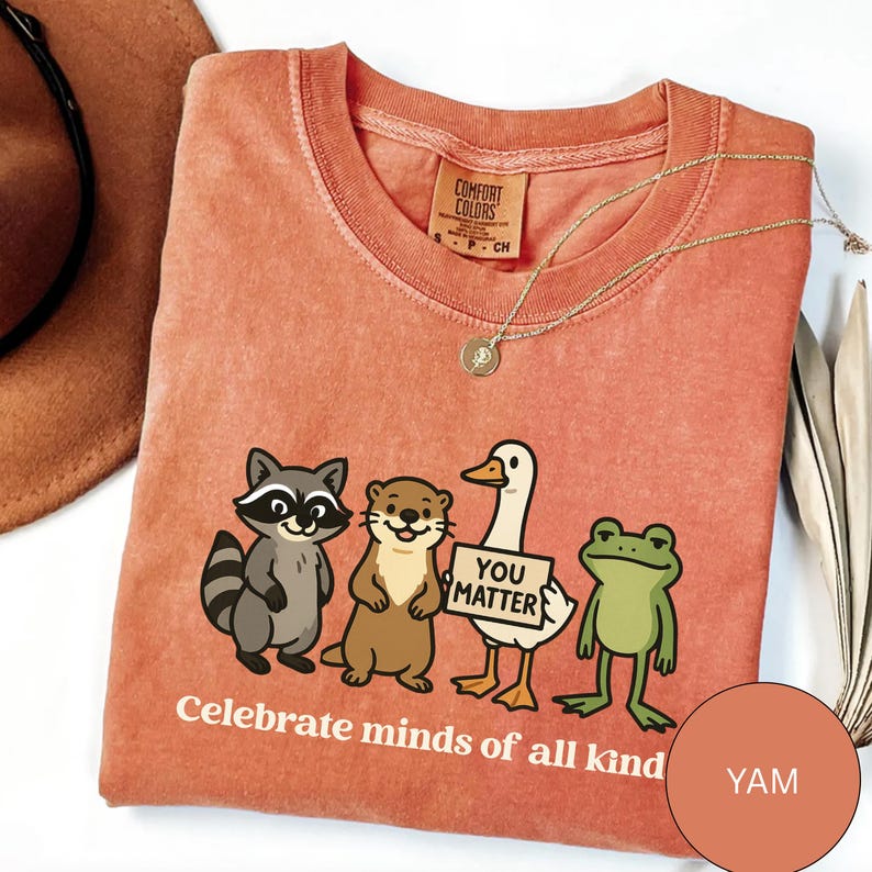 K&ouml;nnte beinhalten: Korallfarbenes T-Shirt mit Rundhalsausschnitt und Grafik eines Waschb&auml;ren, Otters, einer Ente mit einem Schild mit der Aufschrift "You Matter" und einem Frosch. Der Text "Celebrate minds of all kind" steht darunter. Eine goldene Halskette liegt auf dem Shirt.
