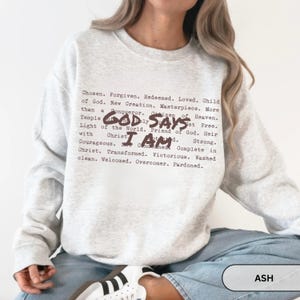 K&ouml;nnte beinhalten: Hellgraues Sweatshirt mit dem Text "GOD SAYS I AM" in dunkelbrauner Schrift. Das Sweatshirt hat auch eine Liste positiver Affirmationen in einer &auml;hnlichen Schriftart. Das Wort "ASH" befindet sich in einem wei&szlig;en Rechteck.