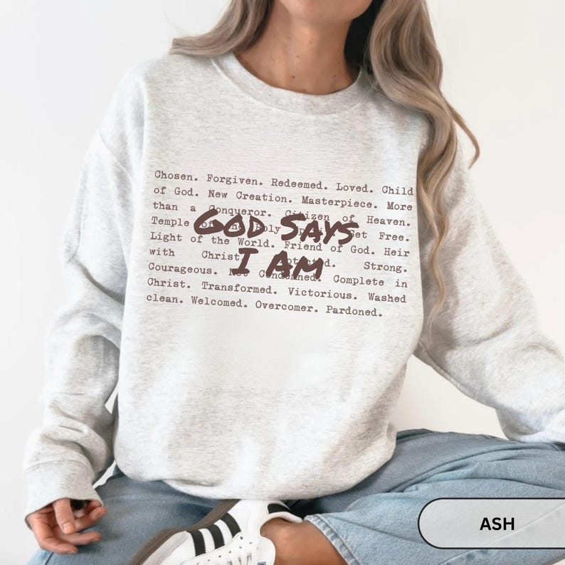 K&ouml;nnte beinhalten: Hellgraues Sweatshirt mit dem Text "GOD SAYS I AM" in dunkelbrauner Schrift. Das Sweatshirt hat auch eine Liste positiver Affirmationen in einer &auml;hnlichen Schriftart. Das Wort "ASH" befindet sich in einem wei&szlig;en Rechteck.