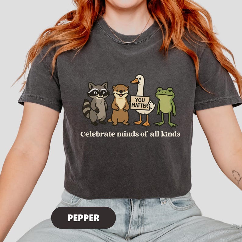 K&ouml;nnte beinhalten: Dunkelgraues T-Shirt mit einer Grafik von Waschb&auml;r, Otter, Ente und Frosch. Die Ente h&auml;lt ein Schild mit der Aufschrift "You Matter". Der Text "Celebrate minds of all kinds" steht darunter. Das T-Shirt ist mit "Pepper" beschriftet.