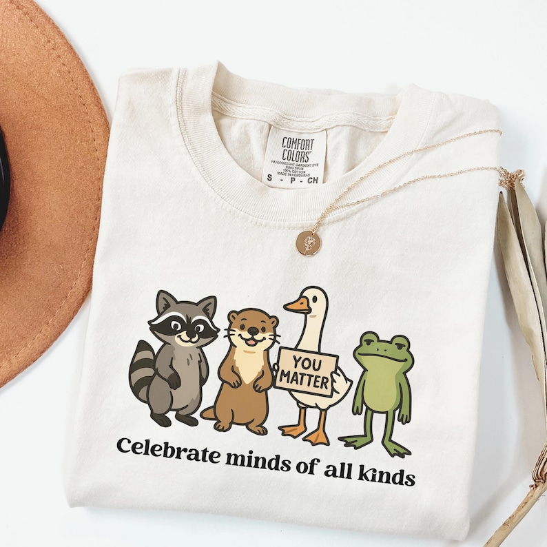 K&ouml;nnte beinhalten: Cremefarbenes T-Shirt mit einem Waschb&auml;ren, einem Otter, einer Ente mit einem "You Matter"-Schild und einem Frosch. Der Text "Celebrate minds of all kinds" ist unter den Zeichentieren aufgedruckt. Eine goldene Halskette liegt &uuml;ber dem Shirt.