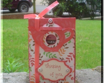 Pode incluir: Uma caixa de presente de chá rosa e branca com um recorte de um bule de chá e as palavras "thinking of you".