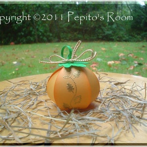 Puede incluir: Una pequeña calabaza de papel naranja y marrón con un tallo verde y una cinta dorada. La calabaza está sentada sobre un lecho de paja.