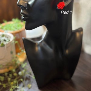 May include: A pair of red crocheted flower earrings. The earrings are shown on a black mannequin head. The text "Red 1" is written below the earrings.