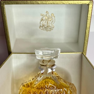 May include: A vintage perfume bottle with a clear, faceted stopper and a gold-colored liquid inside. The bottle is diamond-patterned and sits in an open, gold-trimmed box with a brown lid and a gold emblem inside.