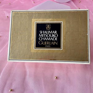 Könnte beinhalten: Eine rechteckige, goldfarbene Schachtel mit strukturiertem Muster und einem schwarz-goldenen Etikett. Auf dem Etikett steht "SHALIMAR MITSOUKO CHAMADE GUERLAIN PARIS". Die Schachtel liegt auf einer rosa Oberfläche mit goldenen Perlen.