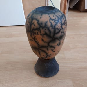 May include: A decorative vase with a unique design. The vase has a rounded body with a dark base and a speckled, orange and black pattern. The top is dark, and the vase sits on a textured base.