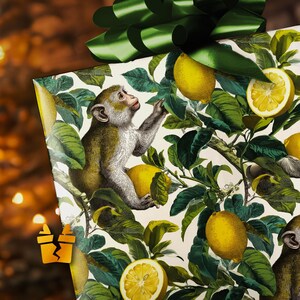 May include: Gift-wrapped present with a green bow. The wrapping paper features a botanical illustration of lemons, leaves, and a monkey. The monkey is reaching for a lemon. The background is blurred with warm tones.