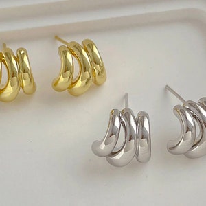 May include: Gold and silver-toned stud earrings. Each earring features three curved, connected segments. The gold earrings have a shiny finish, while the silver earrings have a polished appearance. The earrings are displayed on a white surface.