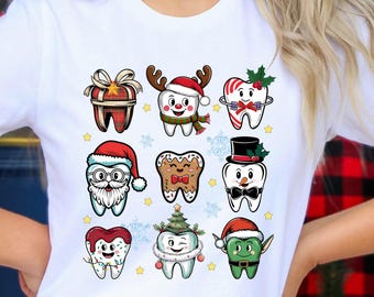 May include: White t-shirt featuring a festive dental-themed design. The graphic includes cartoon teeth dressed in holiday attire, such as Santa hats and reindeer antlers. The design is colorful and playful, perfect for the holiday season.