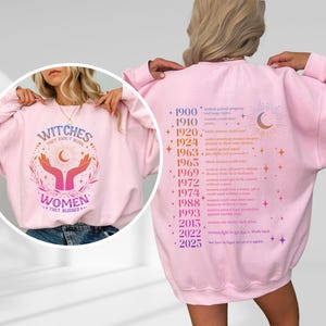 May include: Pink sweatshirt with a graphic print of hands holding a crescent moon and stars. The text "Witches They Don't Burn Women They Burned" is printed on the front. The back of the sweatshirt has a list of historical events related to women's rights.