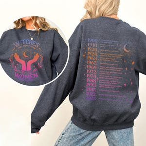 May include: A dark gray sweatshirt with a graphic print on the front that says "Witches, they don't burn, they rise" with a crescent moon and two hands holding a star. The back of the sweatshirt has a list of years and events related to women's rights.