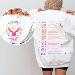 May include: White sweatshirt with a pink and purple graphic of hands holding a crescent moon and stars with the text "Witches they don't burn women they shine" on the front. The back of the sweatshirt has a list of historical events related to women's rights in the United States.