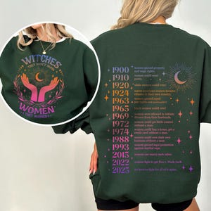 May include: Green sweatshirt with a graphic print of a crescent moon and stars on the back. The front of the sweatshirt has a graphic print of two hands reaching up to a crescent moon with the text "Witches They Don't Burn Women They Rise" in a rainbow gradient.