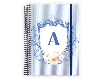 May include: Spiral-bound notebook with a blue and white striped cover. The cover features a blue shield design with the letter "A" in the center, surrounded by floral accents. A blue elastic band secures the notebook.
