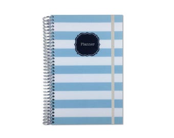 May include: A spiral-bound planner featuring a light blue and white horizontal striped pattern. The cover includes a centered dark navy blue scalloped label with the word Planner printed in white text. A thin vertical elastic band secures the cover.