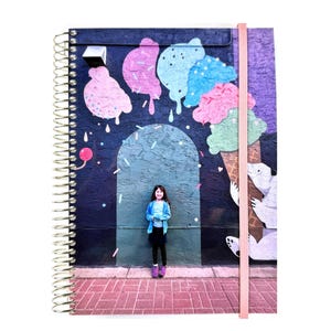 May include: Spiral-bound notebook featuring a colorful mural of ice cream cones and a girl standing in front of it. The notebook has a pink elastic band closure and a gold spiral binding. The cover art includes pink, blue, and green ice cream.
