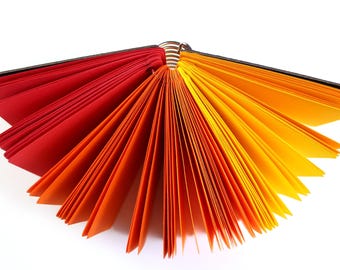 May include: An open notebook with pages fanned out in a gradient of colors. The pages transition from red to orange to yellow, creating a vibrant and eye-catching display. The binding is dark, and the pages are on a white background.